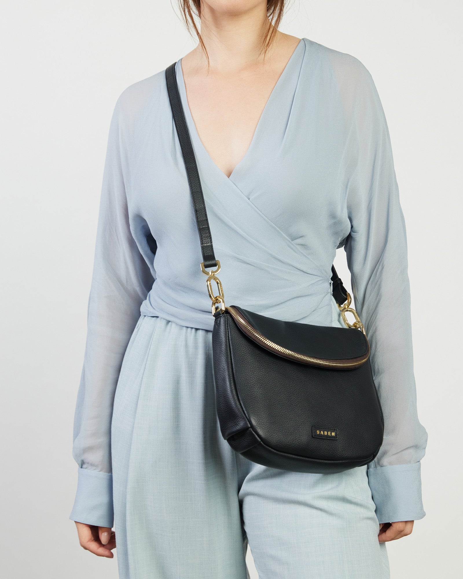 A black leather crossbody bag with a functional zipped flap closure and a detachable strap, featuring gold-plated hardware.