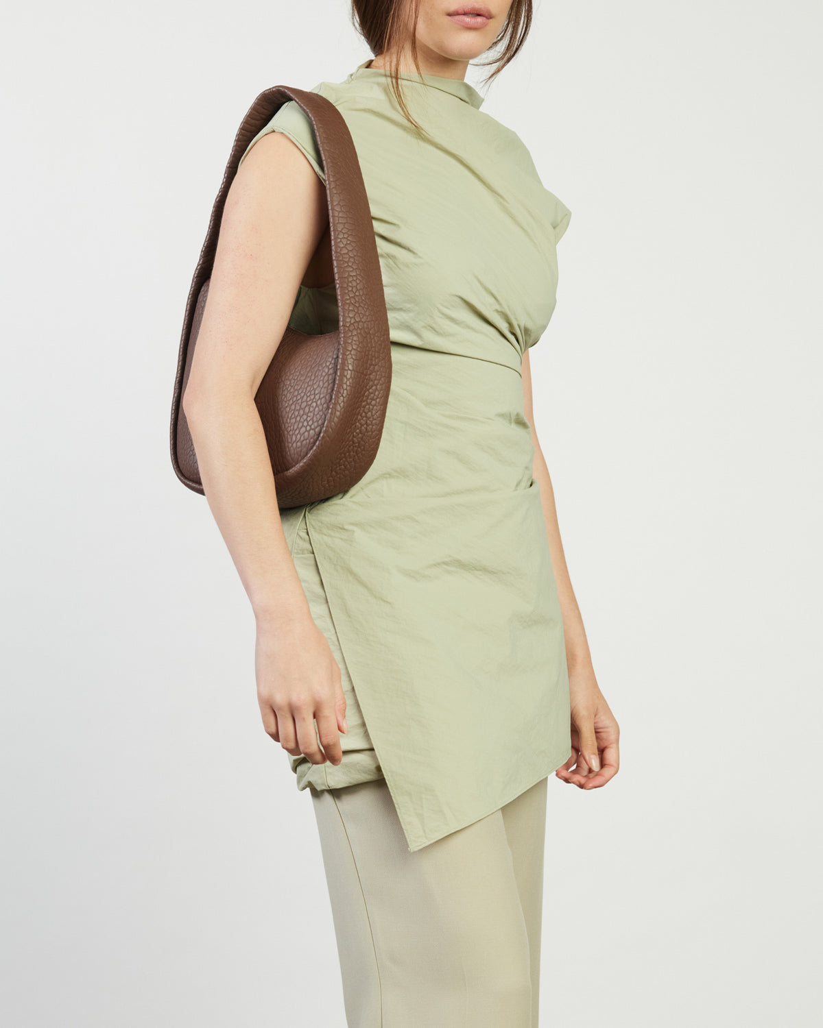Woman wearing a light green sleeveless top and beige pants, holding a brown leather bag on a white background