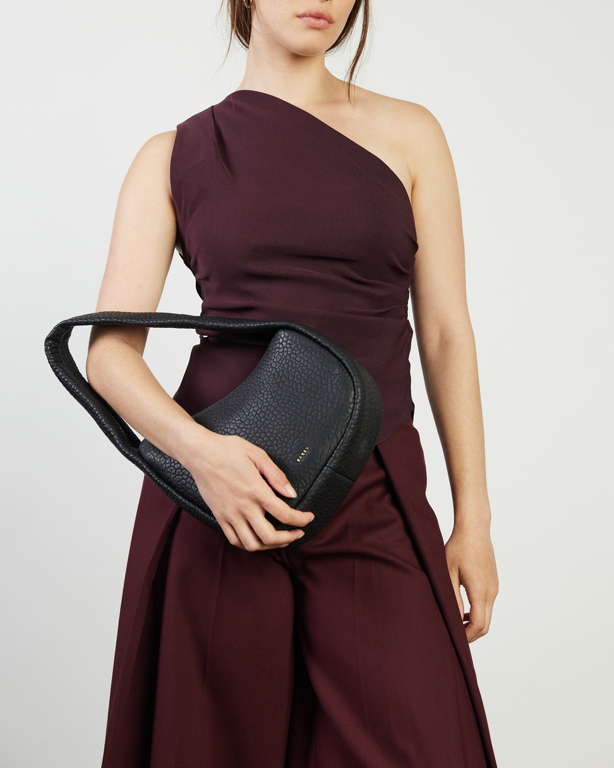 Woman holding a black handbag wearing a burgundy one-shoulder dress on a light gray background