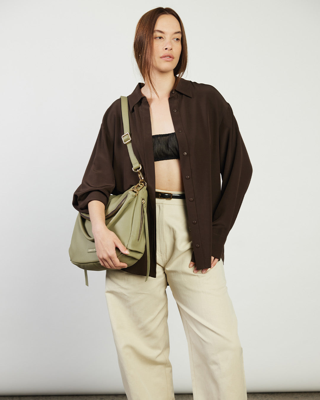 Person wearing a brown shirt, black top, beige pants, and holding a green bag against a white background