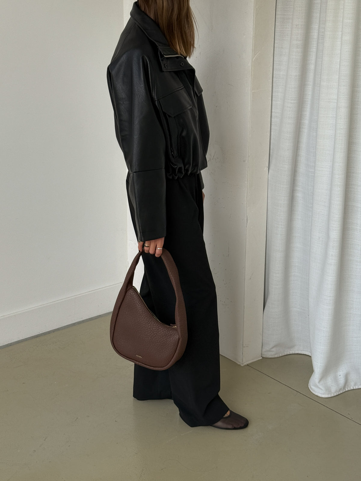 Person wearing a black outfit holding a brown handbag in a minimalistic setting.