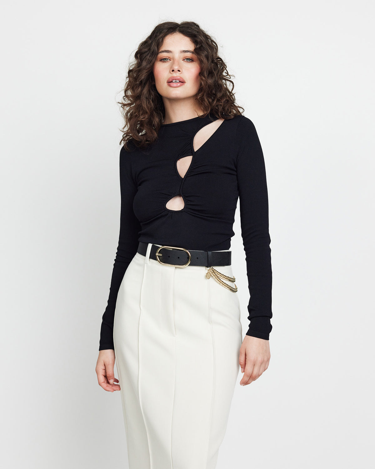 Woman wearing a black long-sleeve top with cut-out design and white pants on a white background