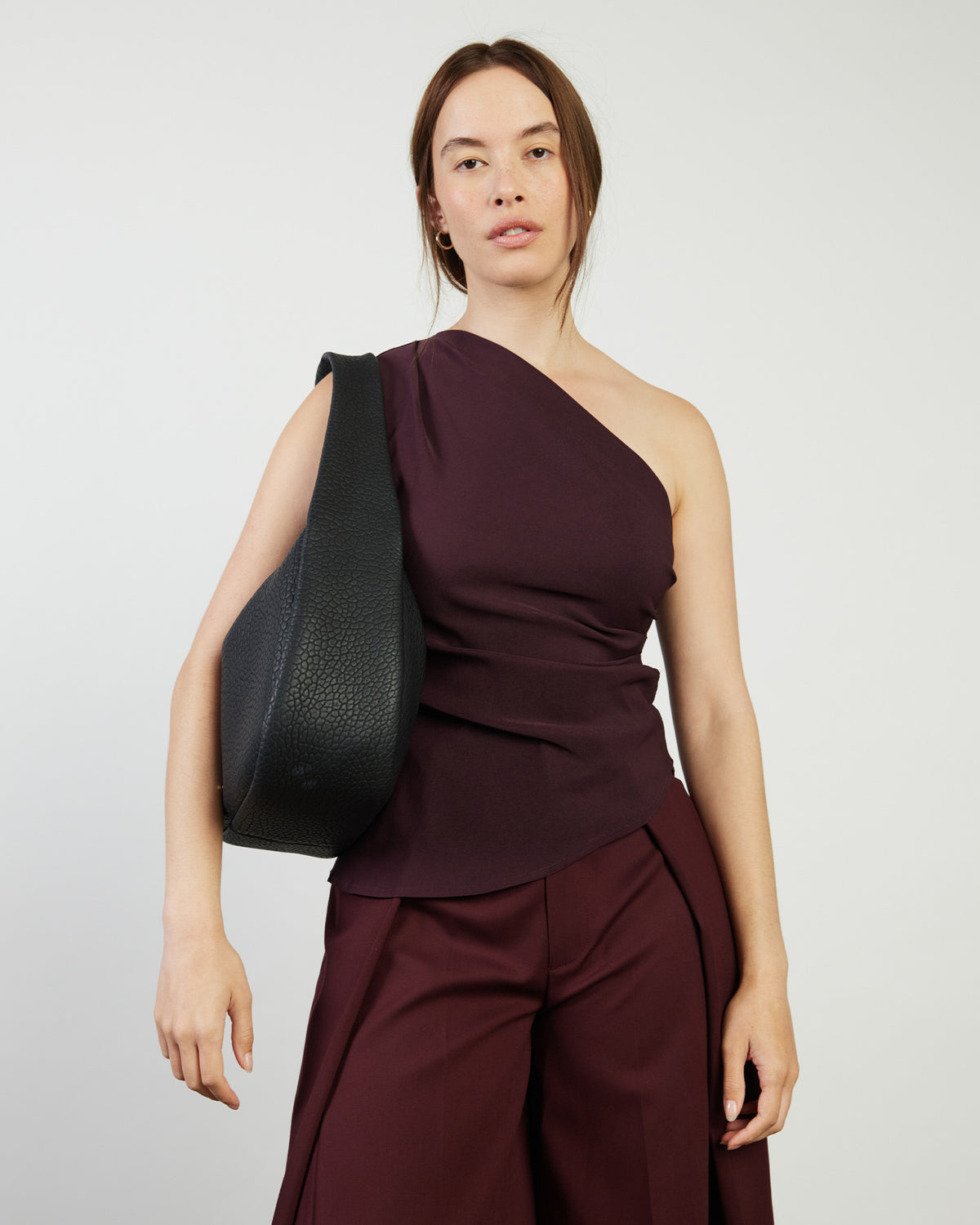 Woman wearing a burgundy one-shoulder top and pants with a black bag on a white background