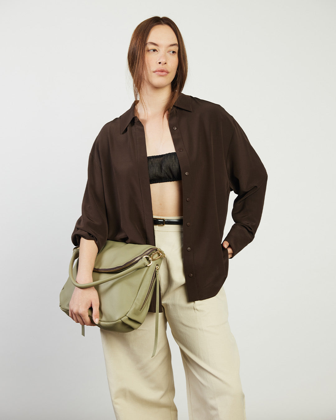Person wearing a brown shirt, black top, beige pants, and holding a green bag against a white background