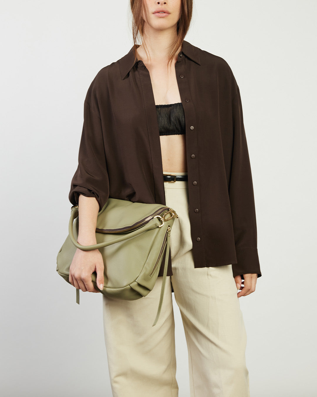 Person wearing a brown shirt, black top, beige pants, and holding a green bag against a white background
