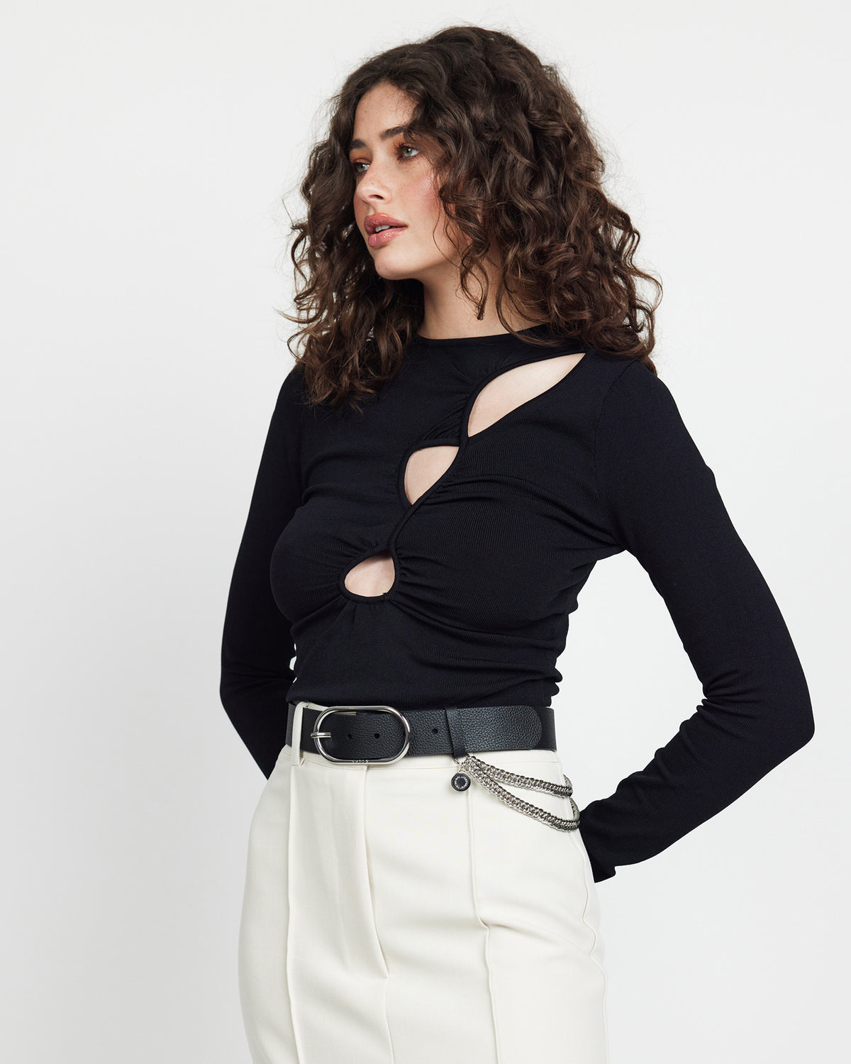 Woman wearing a black long-sleeve top with cut-out design and white pants on a light background
